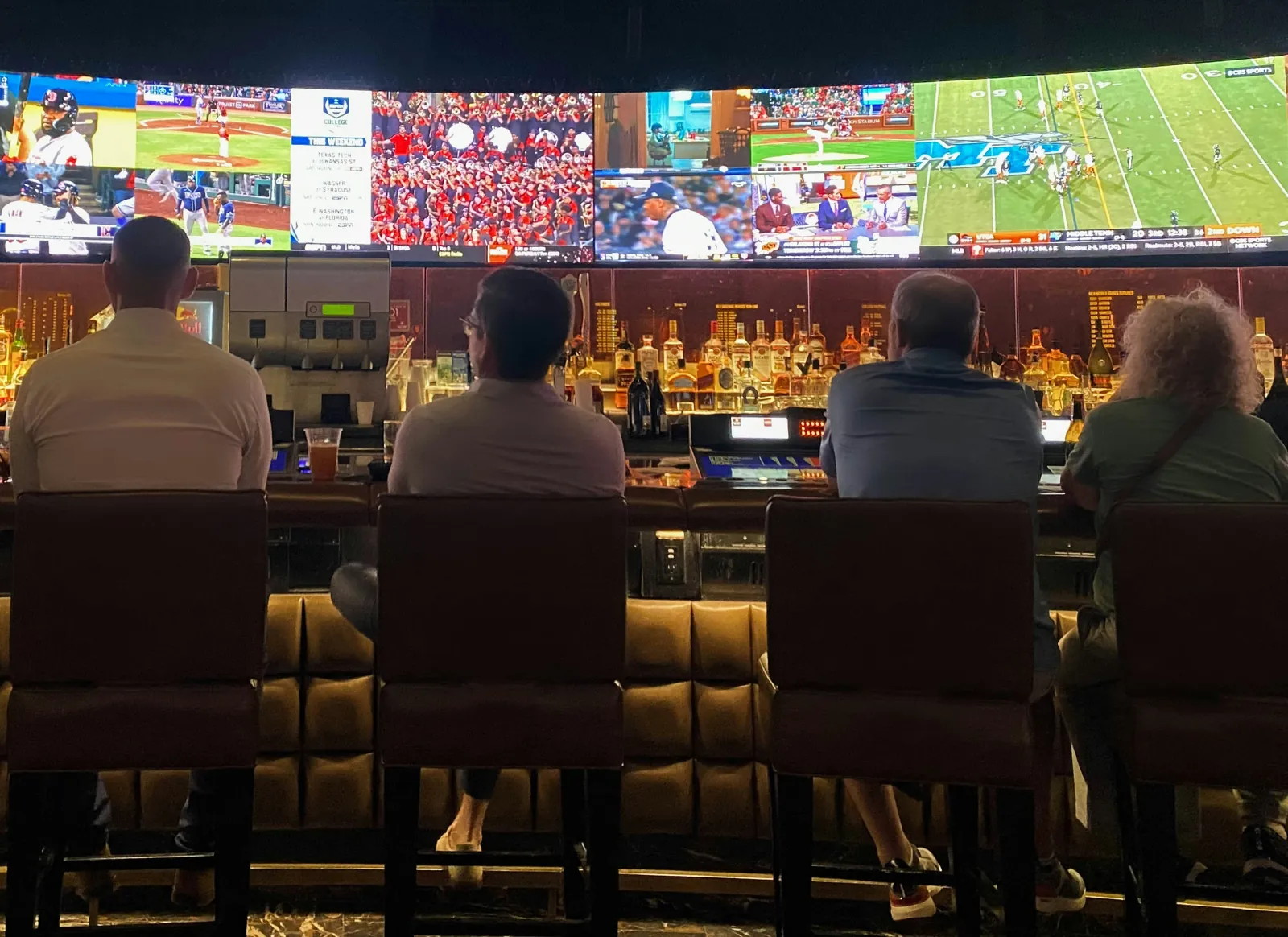 Patrons seated at a bar, seen from behind, facing a curved video wall showing sports